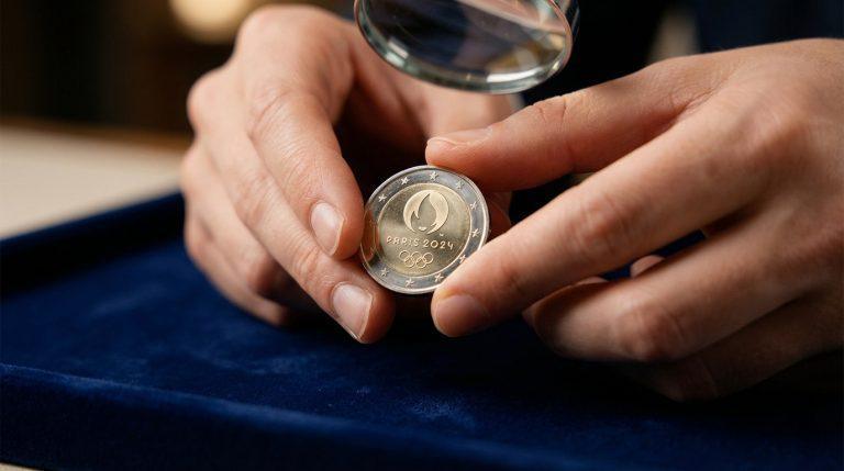 Gros plan sur des mains examinant à la loupe une pièce de 2 euros commémorative des JO Paris 2024 sur velours bleu.